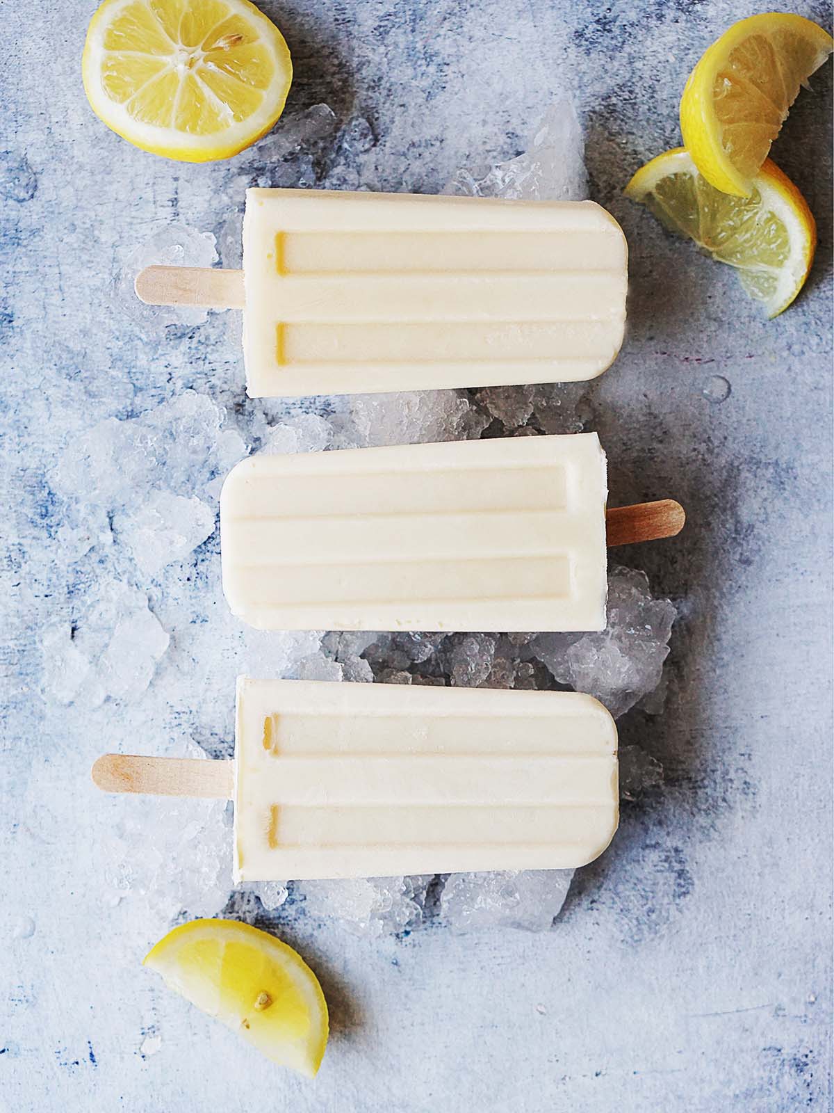 Three lemon paletas on a blue background with ice and lemon wedges.
