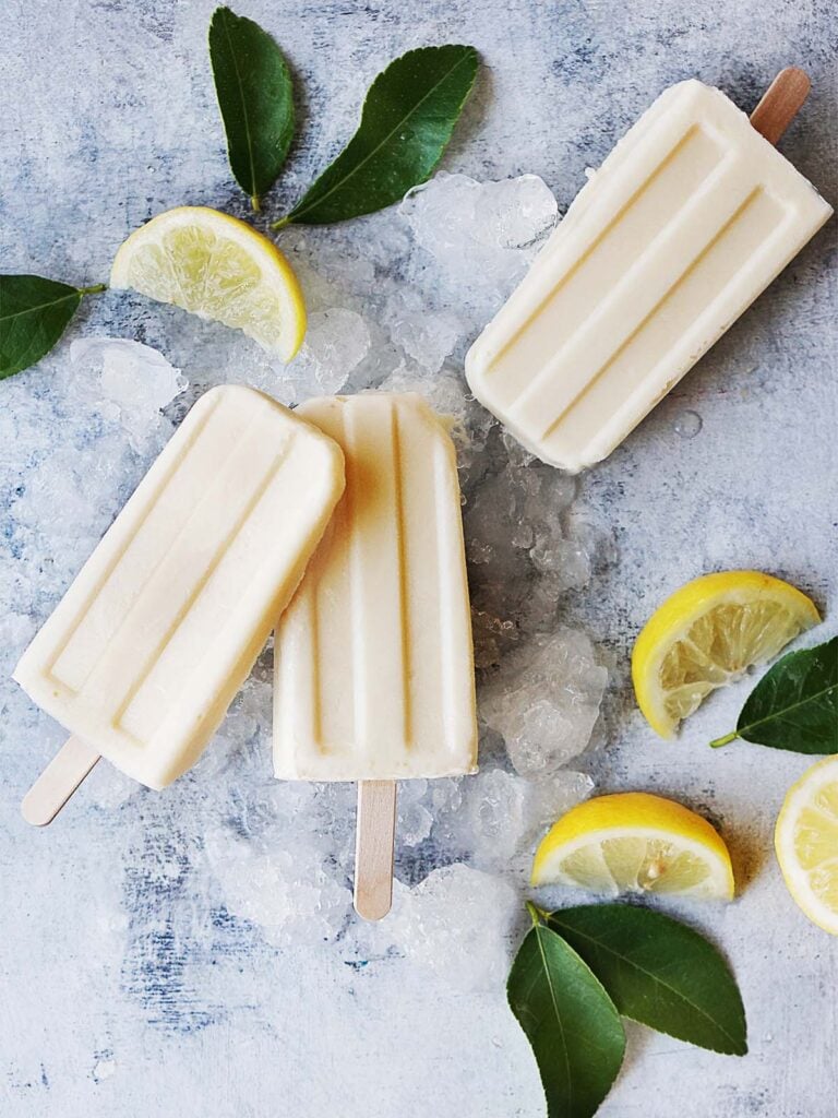 Three lemon popsicles on a blue background with ice and lemon wedges.