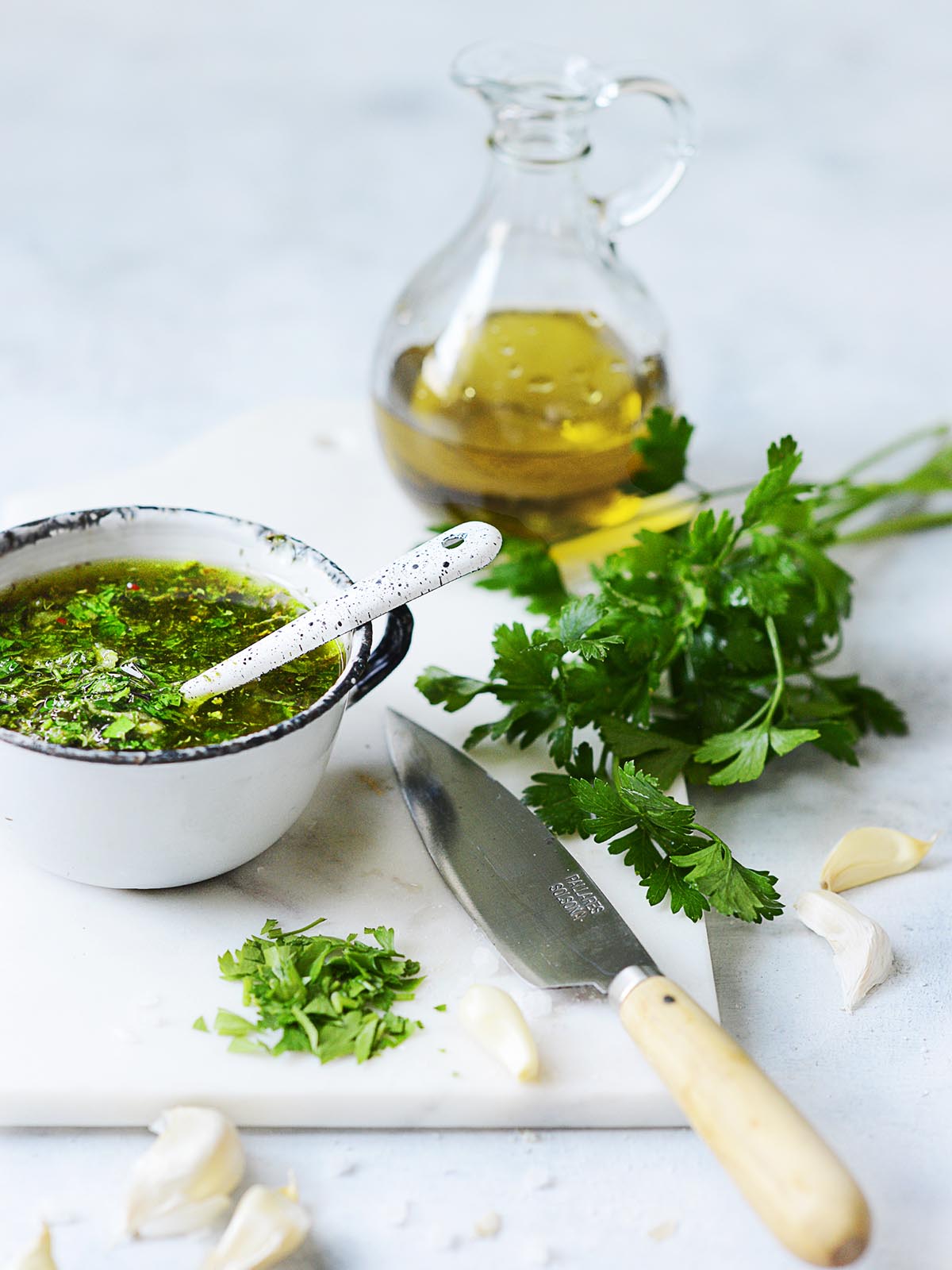 A white vintage cup with the green sauce, fresh parsley on the side and an olive oil jar in the background.