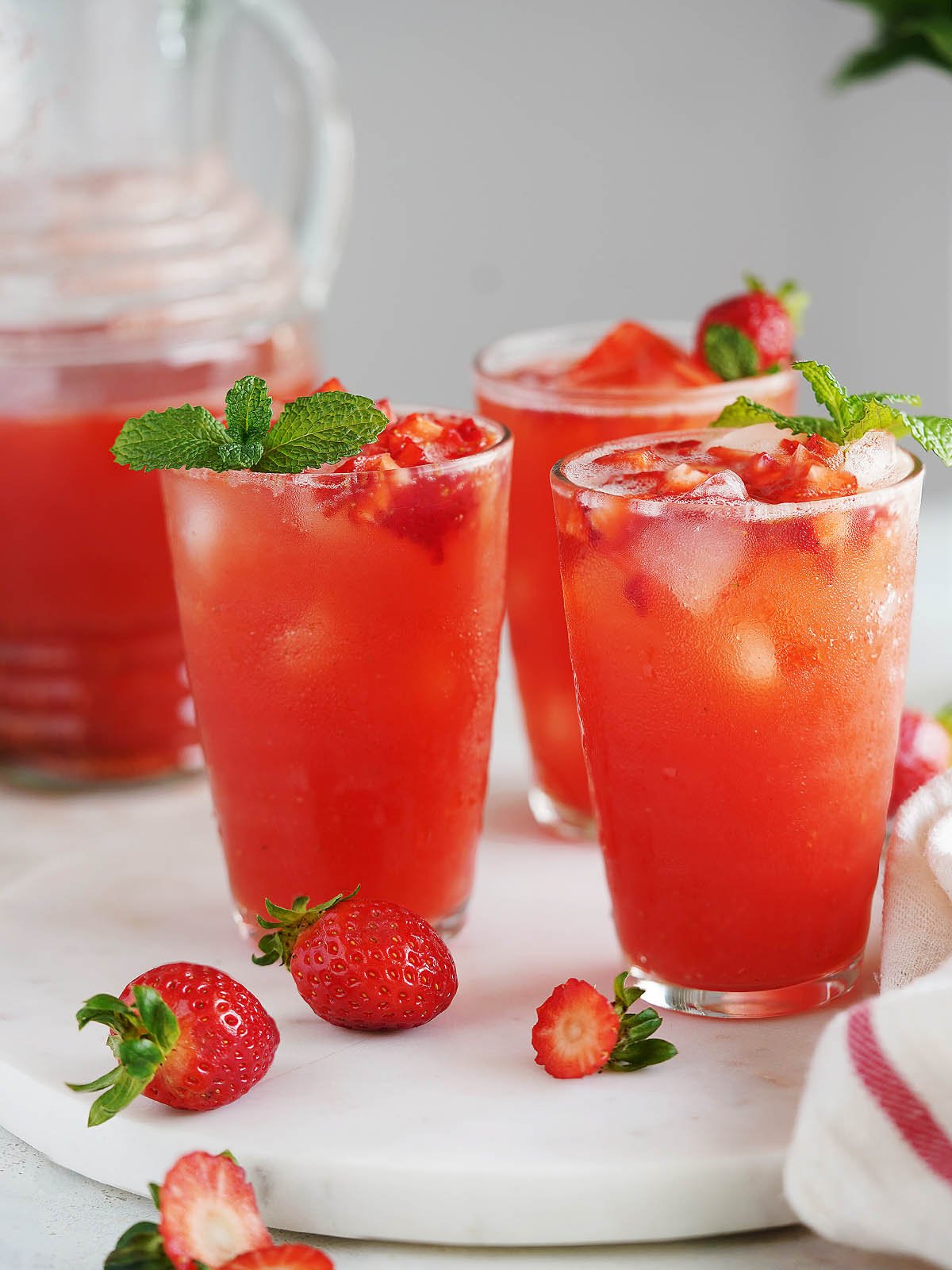 Three glasses with ice and strawberry water topped with small chunks of strawberries and the jar in the background.