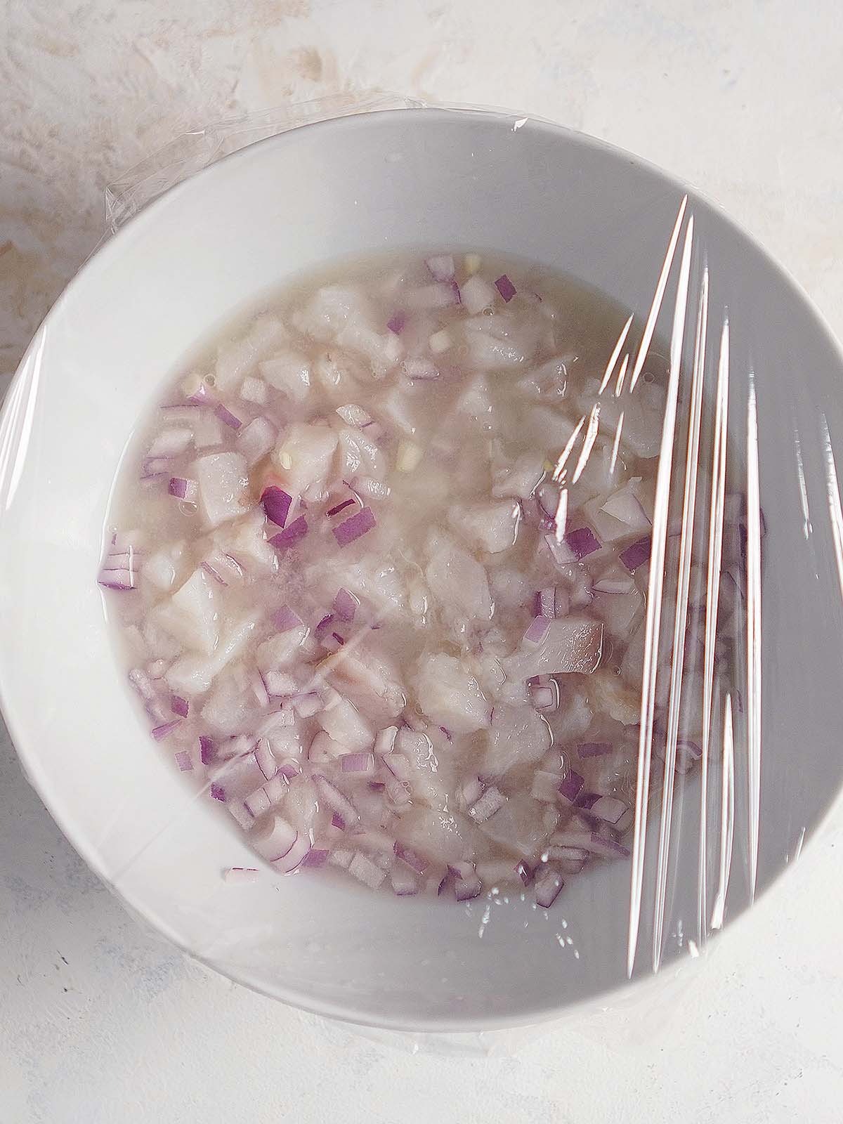 A white bowl with cubed fish, chopped red onions and lime juice covered with plastic wrap.