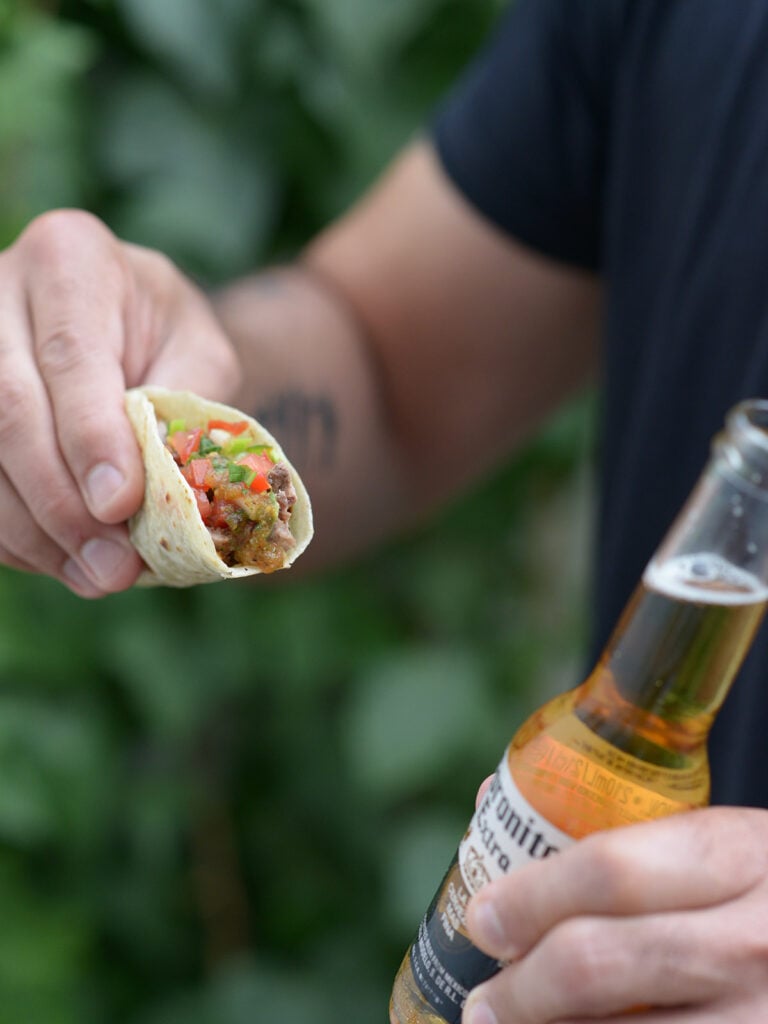 A hand holding a carne asada taco with a beer on the other hand.
