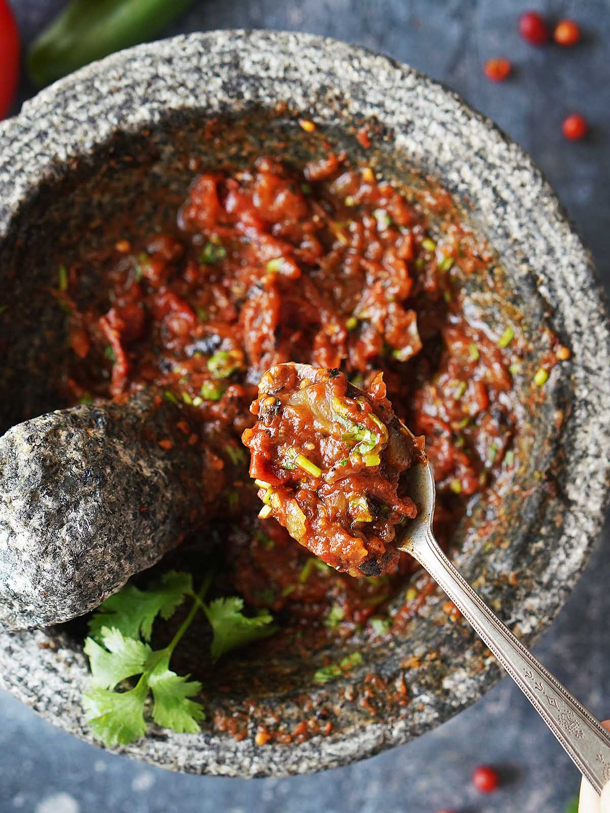 A spoon with chunky salsa with roasted vegetables in a molcajete.