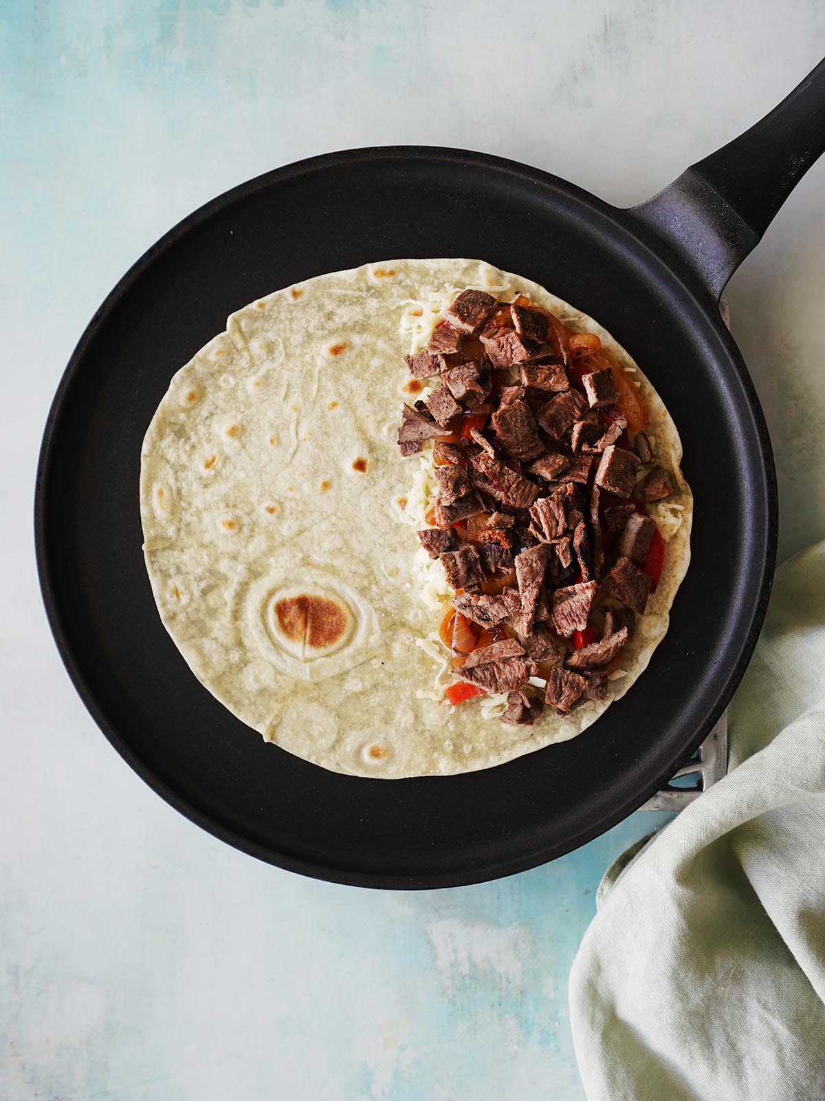 A flour tortilla topped with cheese, sauteed veggies, chopped steak on a skillet.