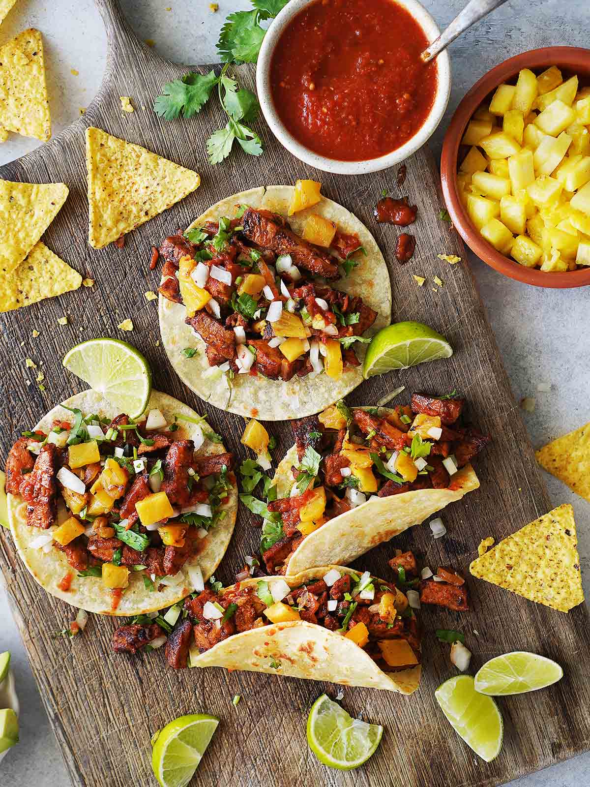 Four tacos al pastor on a wood board with salsa and pineapples on the side.