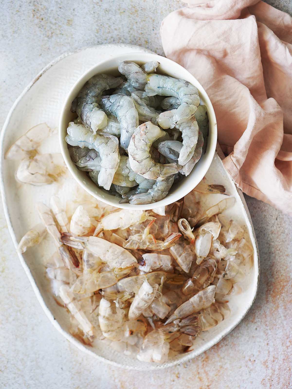Raw peeled shrimp with the peel on the side on a white oval plate.
