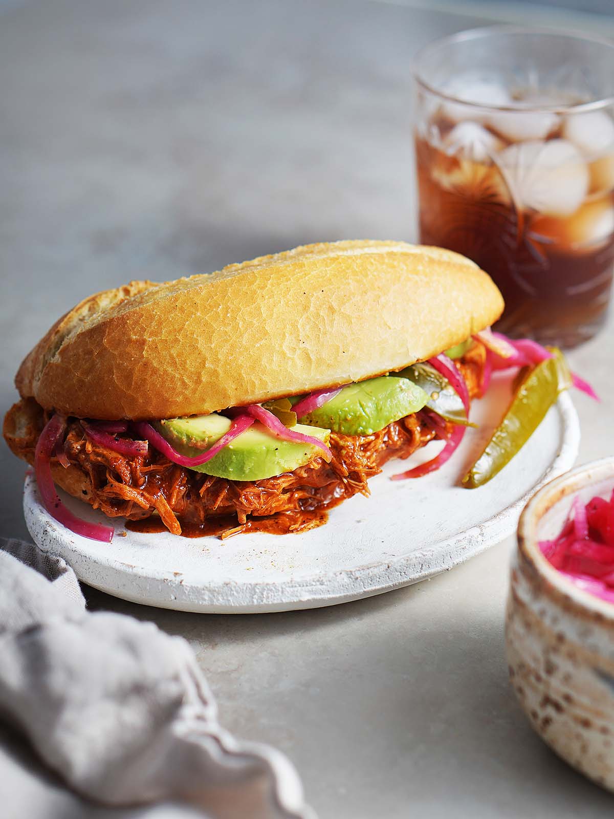 A Cochinita (Pork) Pibil Torta placed on a white plate with a drink on the back.
