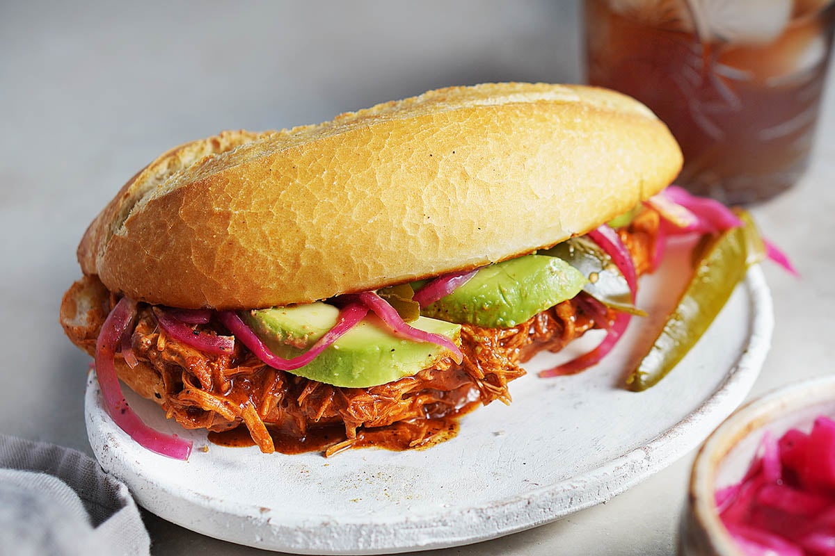 A Cochinita (Pork) Pibil Torta placed on a white plate with a drink on the back.