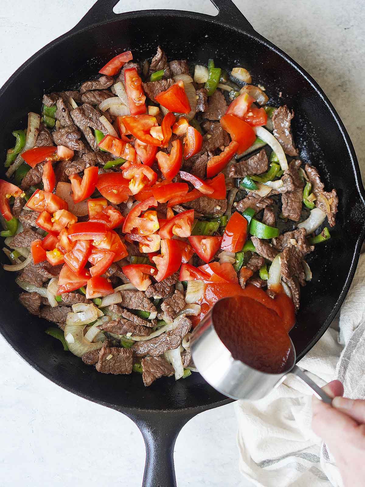 A skillet with cooked beef, onions, chiles & tomatoes and adding tomato sauce to it.