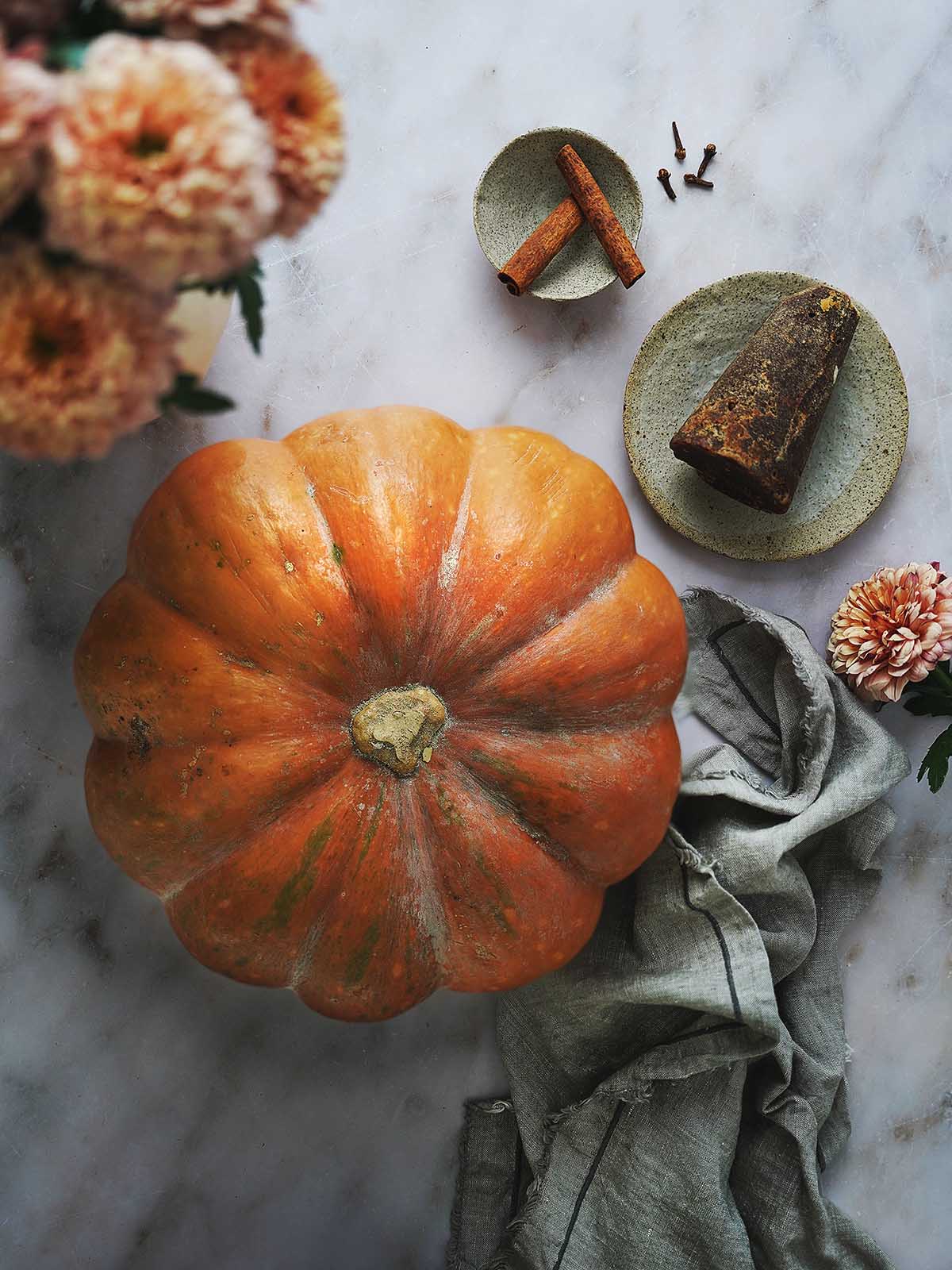A large orange pumpkin, a piloncillo, two cinnamon sticks and 4 cloves on a white background.
