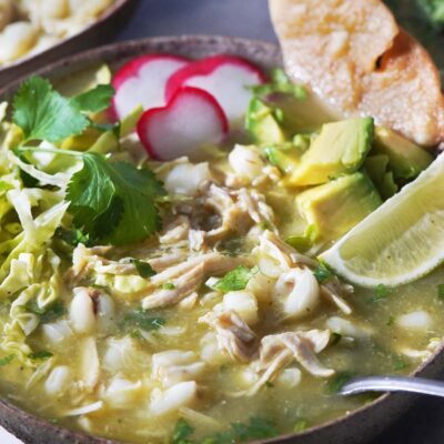 A bowl with pozole, and radishes, avocado and lime as garnish.