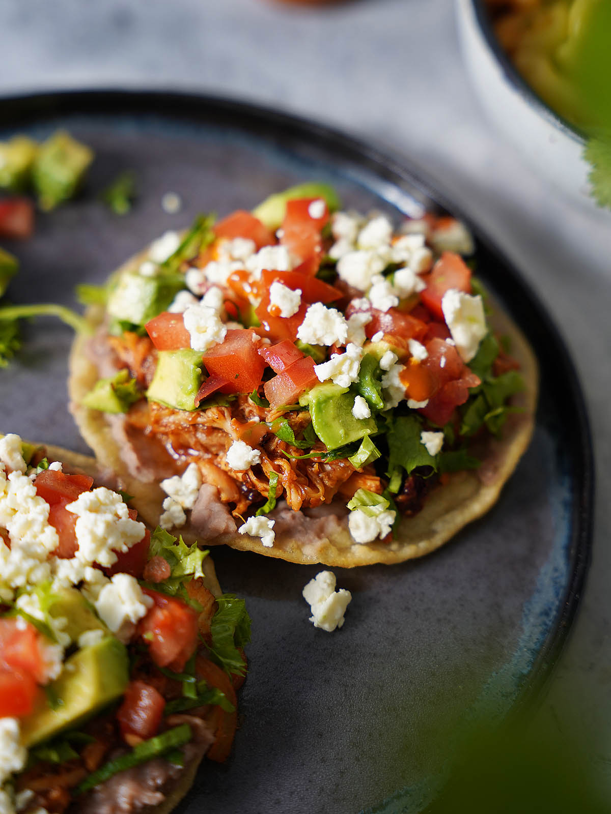 Two chicken tostadas on a plate topped with beans, shredded chicken, tomatoes, avocados and lettuce.