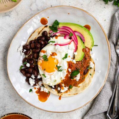 A plate with a huevos rancheros, black beans, avocado and pickled onions.
