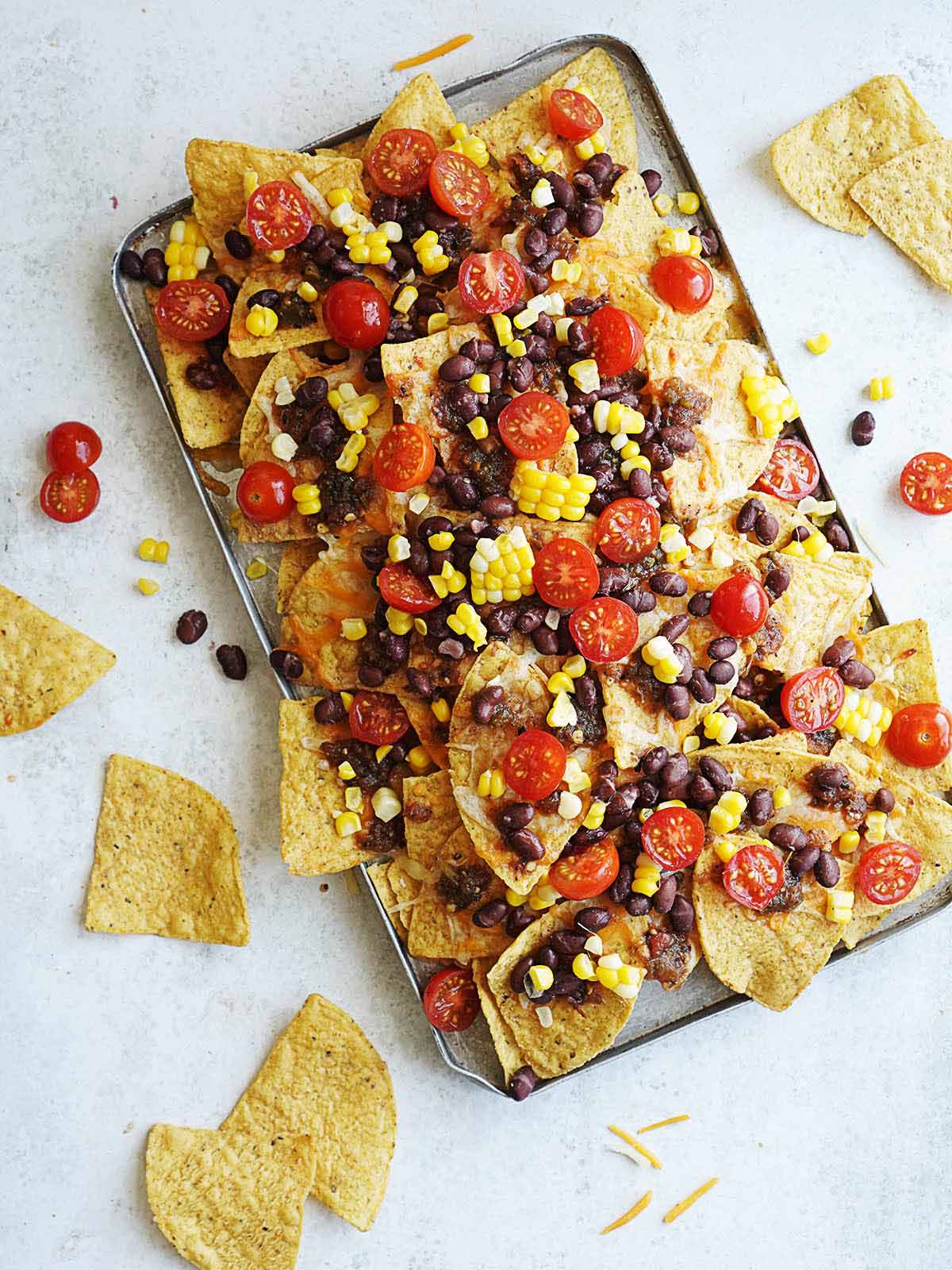 A baking tray with tortilla chips baked with melted cheese topped with tomatoes, black beans & corn kernels.