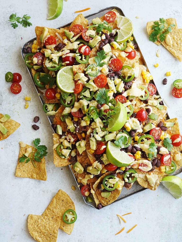 A tray of nachos loaded with chips covered in melted cheese, avocado, tomatoes, black beans, corn, and cilantro.