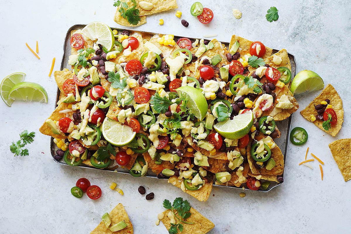 A tray of nachos loaded with chips covered in melted cheese, avocado, tomatoes, black beans, corn, and cilantro.