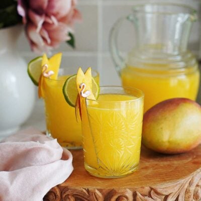 A glass of Agua De Mango in a glass with mango slices as garnish with a jar in the background.