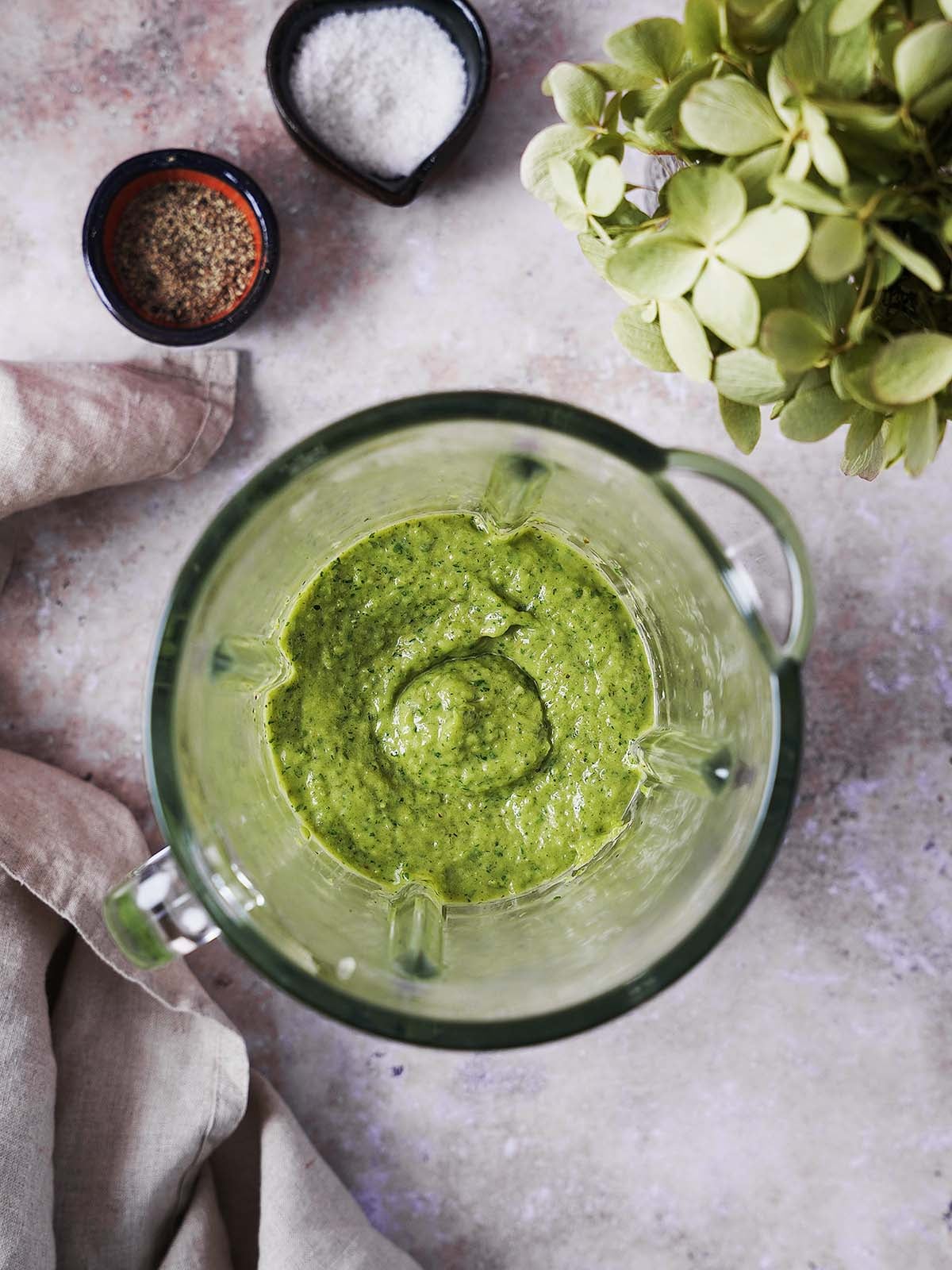 Top view of a blender filled with freshly blended green sauce made from roasted peppers, cilantro, and avocado, with small bowls of salt and pepper on the counter nearby.