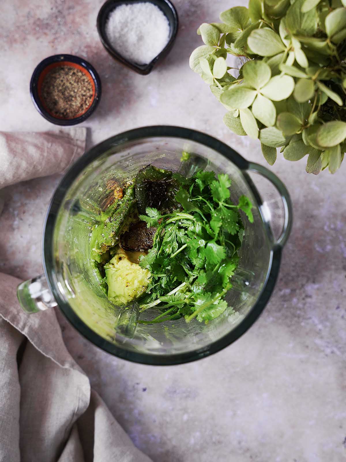 Top view of a blender filled with roasted peppers, fresh cilantro, avocado, and seasonings, ready to be blended into a sauce, with bowls of salt and pepper nearby on a textured surface.