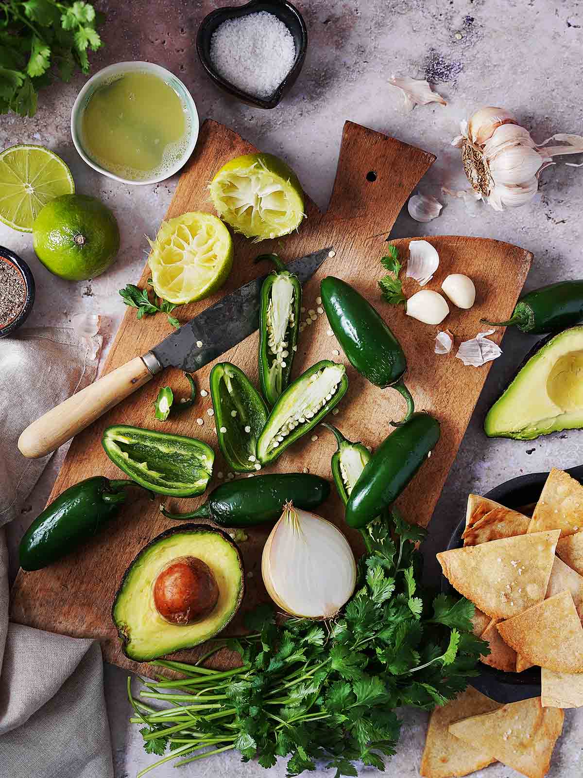 Fresh guacamole ingredients on a wooden cutting board, including halved jalapeños, squeezed limes, avocado, onion, garlic, and cilantro, surrounded by tortilla chips and seasonings.