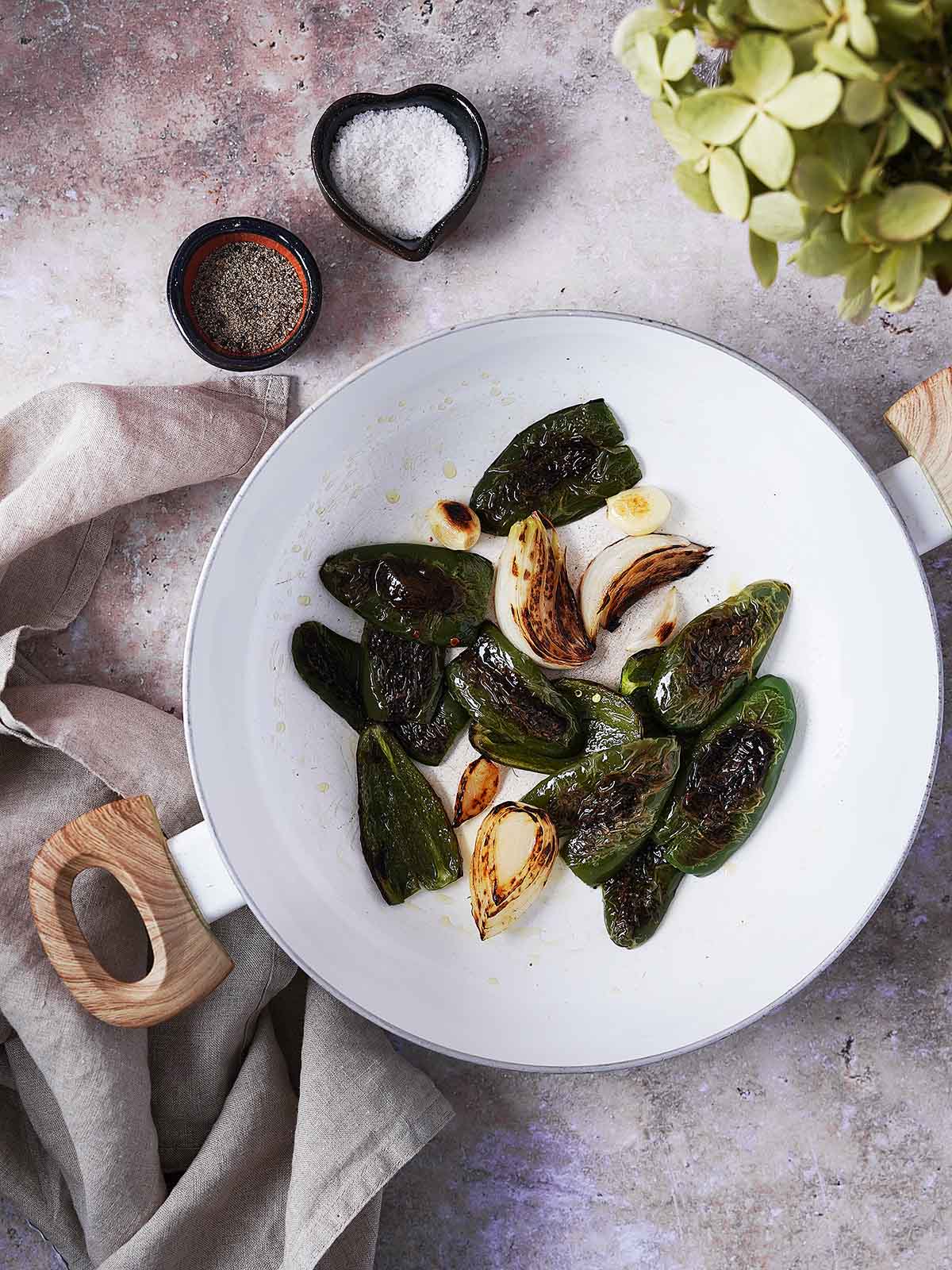 Charred jalapeños, garlic, and onion are roasting in a white skillet, surrounded by small bowls of salt and pepper and a neutral linen cloth on a textured surface.