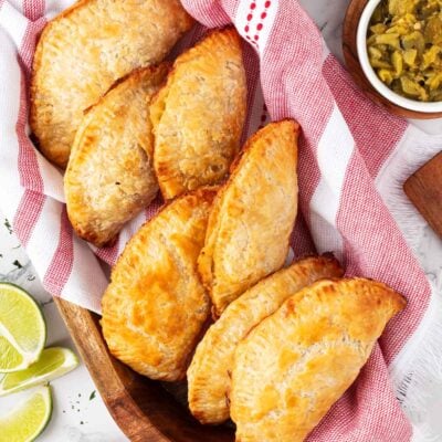 Baked empanadas in a basket lined with a pink kitchen towel with fresh lime wedges and Mexican salsa.