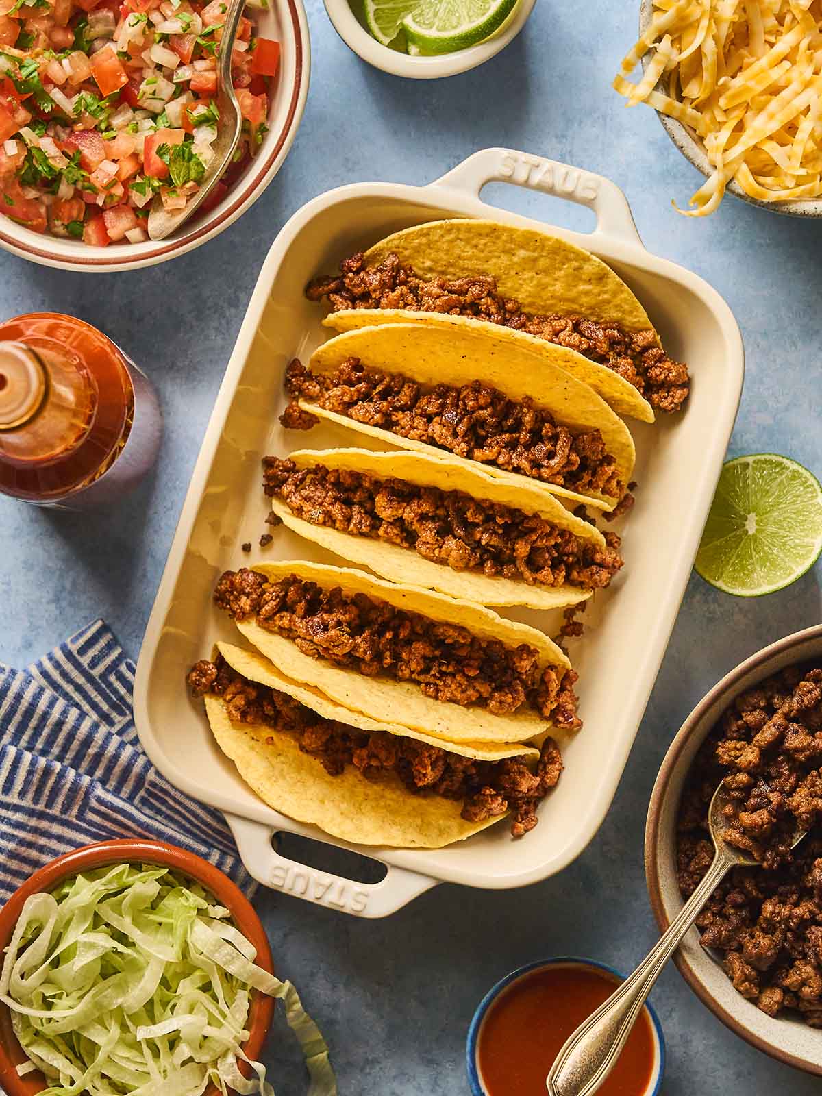 Hard taco shells filled with cooked ground beef in a baking dish surrounded by all fillings in bowls.