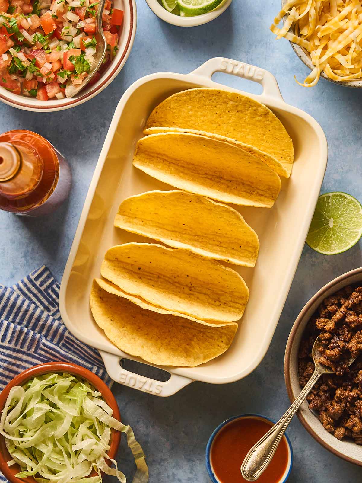 Hard taco shells in a baking dish surrounded by all fillings in bowls.