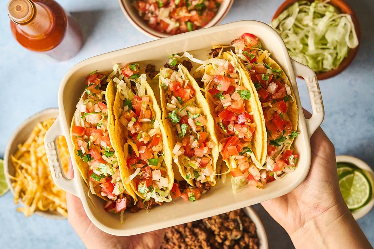 Two hands holding a baking dish filled with ground beef tacos topped with lettuce and salsa fresca.