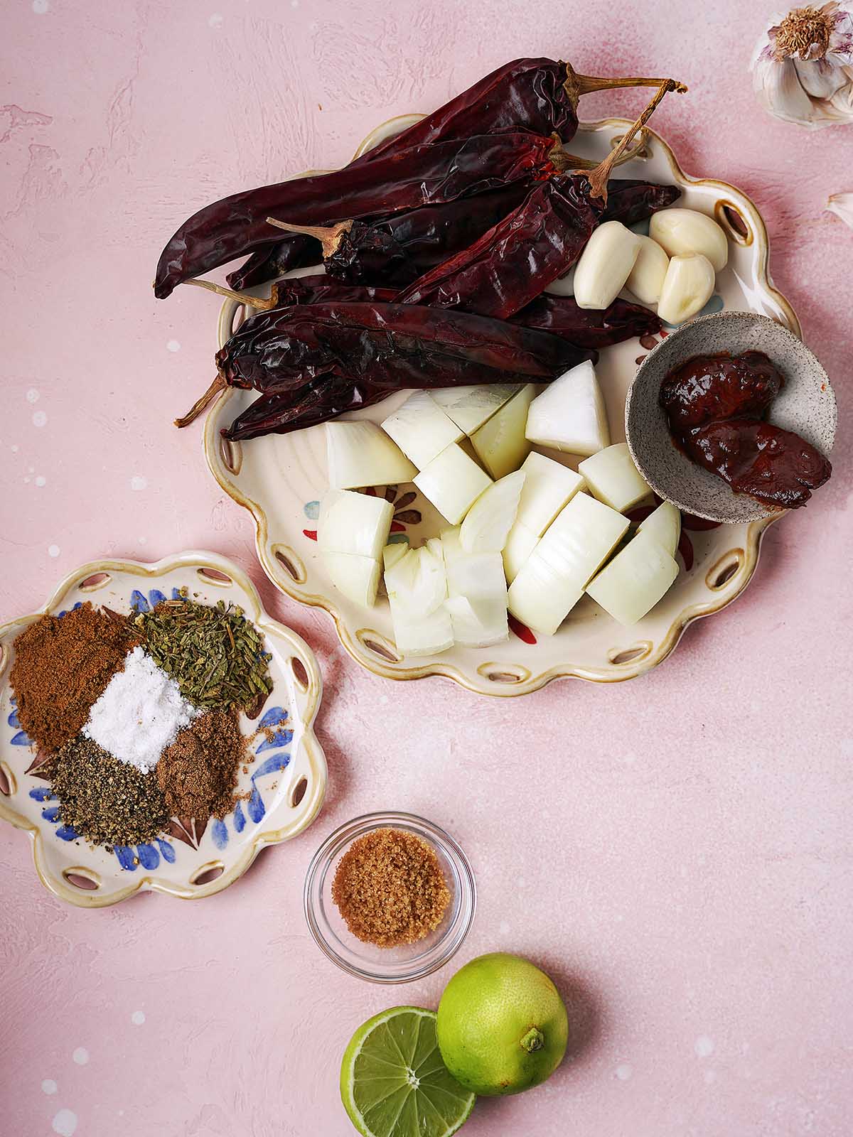 Assorted dried chilies, garlic cloves, chopped white onion, whole spices, salt, dried herbs, a small bowl of ground spice, a halved lime, and a few small dried peppers arranged on decorative plates and bowls on a pink surface.