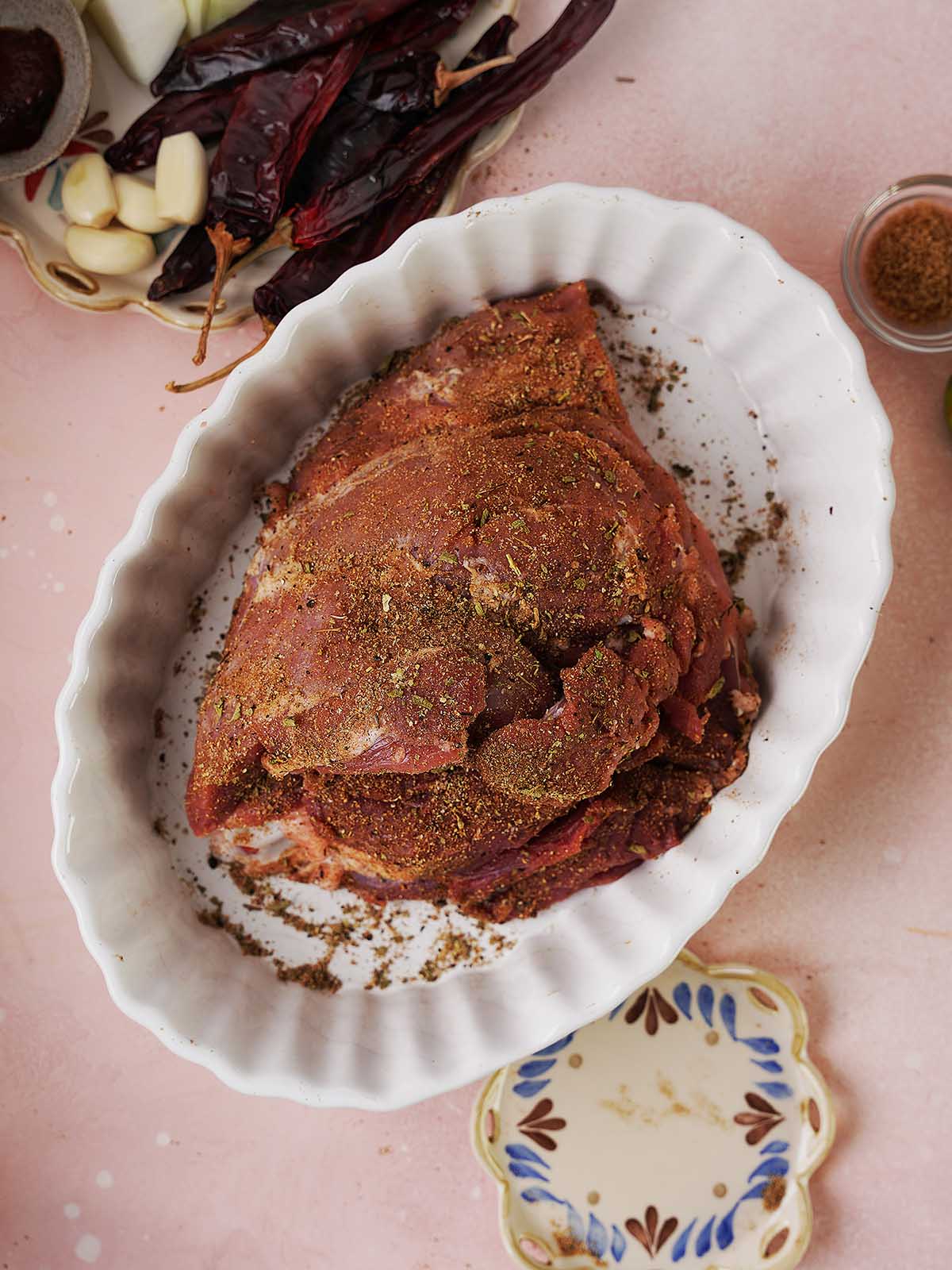 A seasoned piece of meat sits in a white oval baking dish. Surrounding it are dried chiles, peeled garlic cloves, and a small bowl of ground spices, all arranged on a light pink surface.