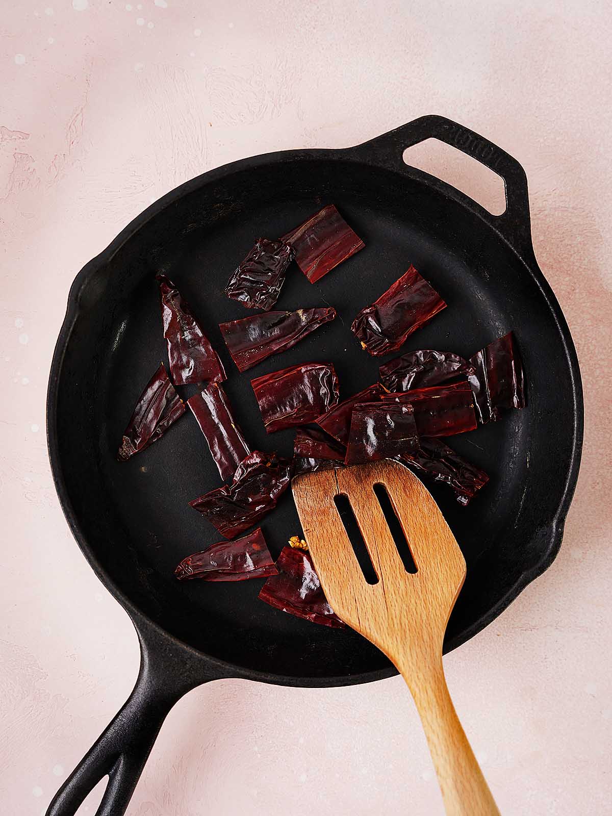A cast iron skillet with dried red chili peppers being stirred by a wooden spatula, set on a light pink textured surface.