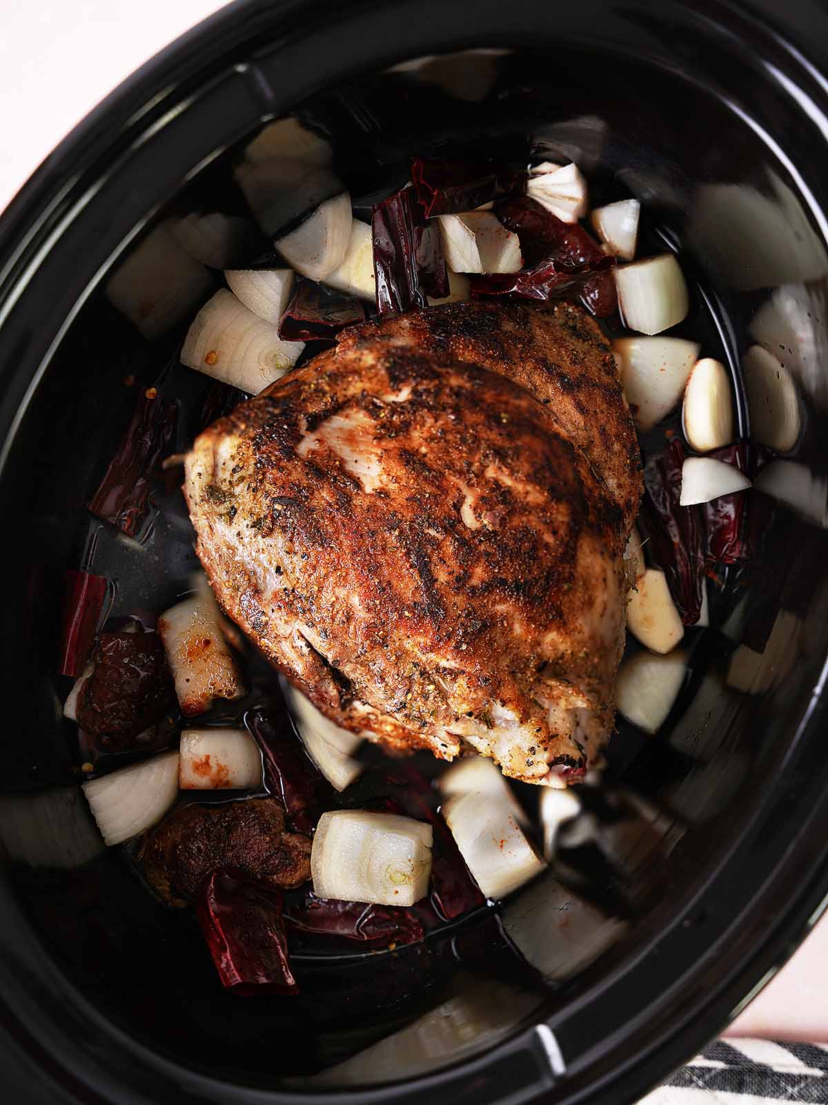 A seasoned pork roast sits in a slow cooker with chopped onions and dried red chilies scattered around it, ready to be cooked.