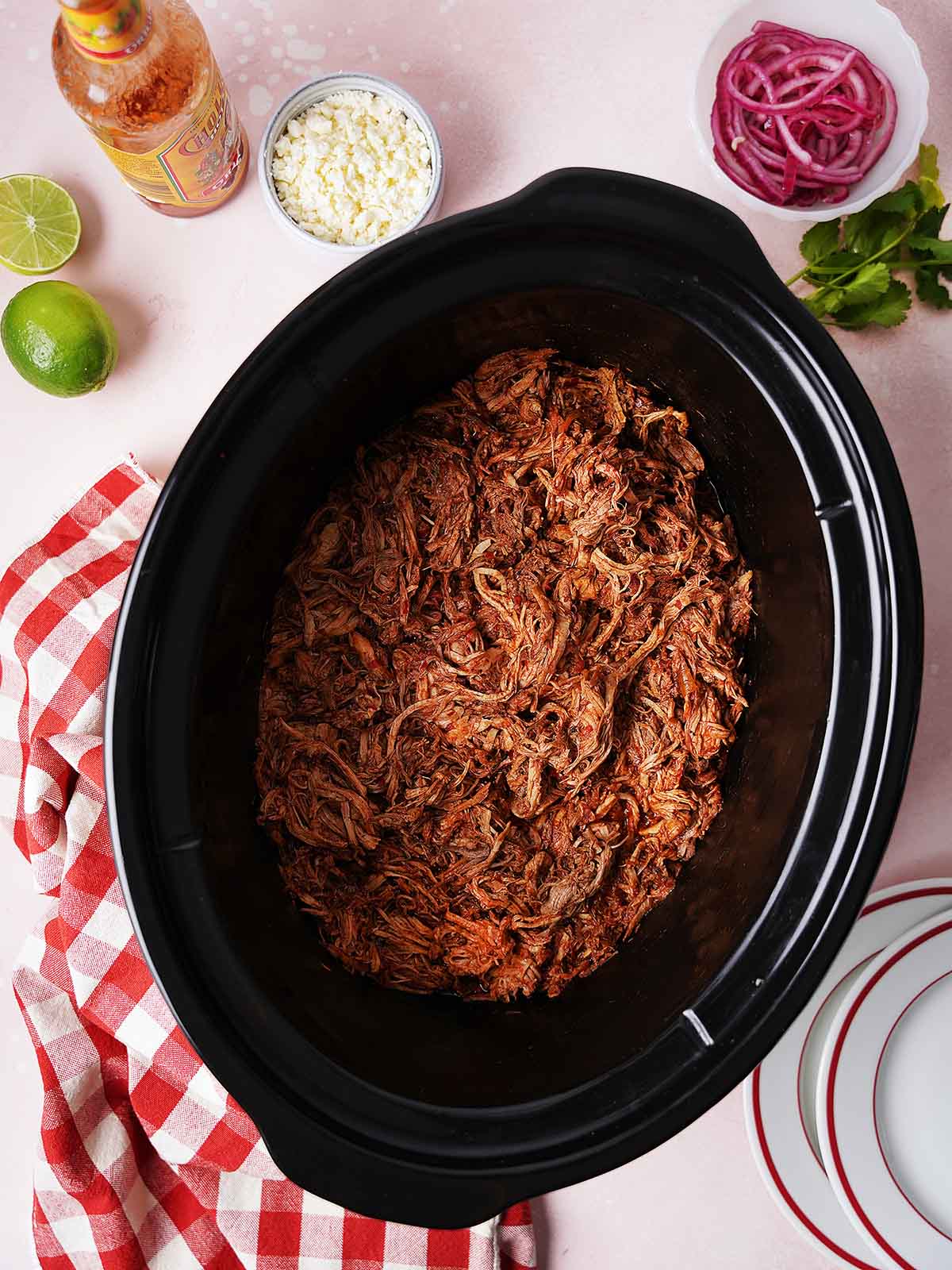 Shredded porkin a slow cooker, surrounded by a red checkered napkin, lime halves, a bottle of hot sauce, a bowl of cheese, pickled onions, and fresh cilantro on a light pink surface.