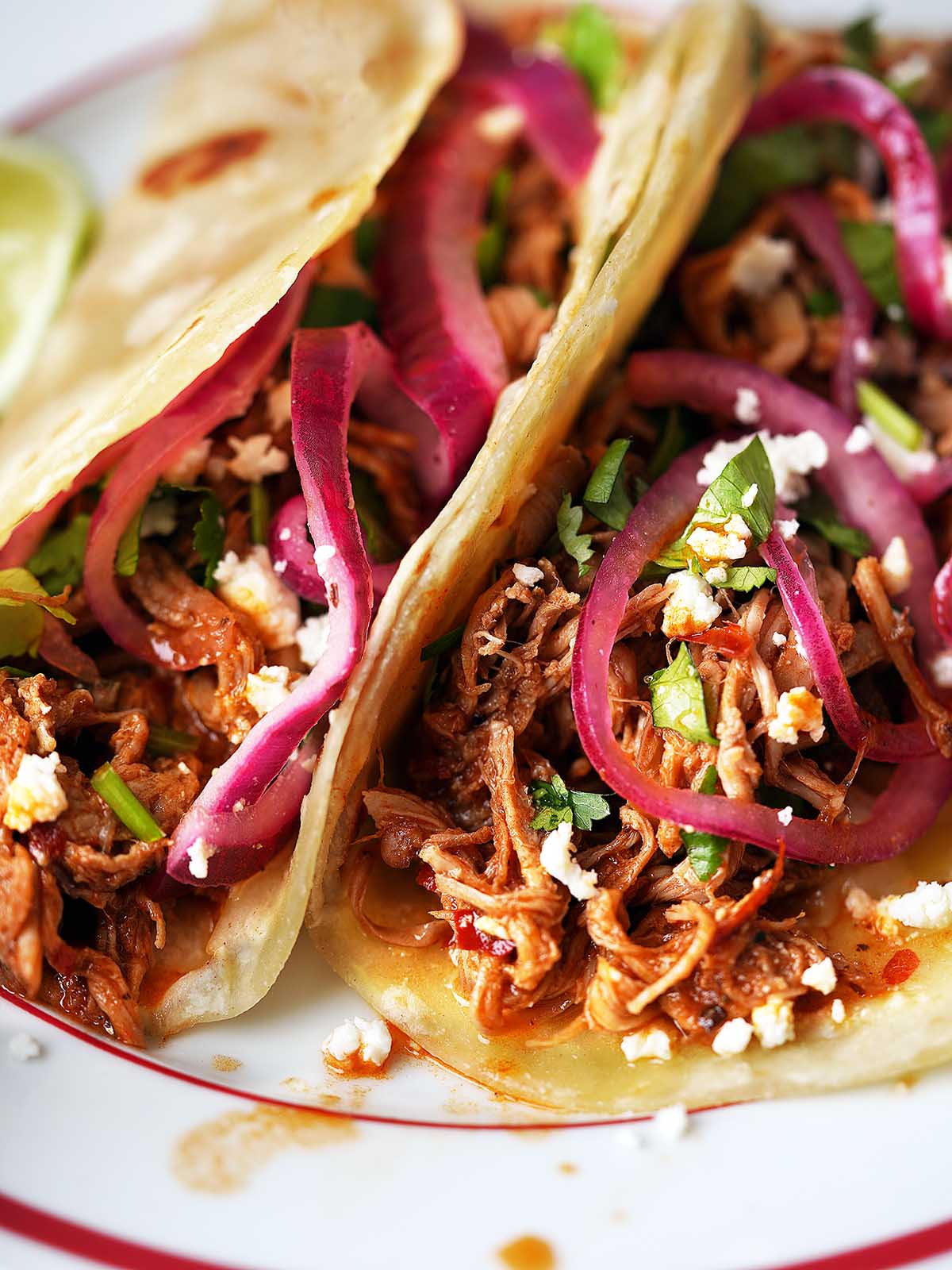 Two pulled pork tacos filled with shredded meat, pickled red onions, cilantro, and crumbled cheese, served on a white plate with a red rim. A lime wedge is partially visible in the background.