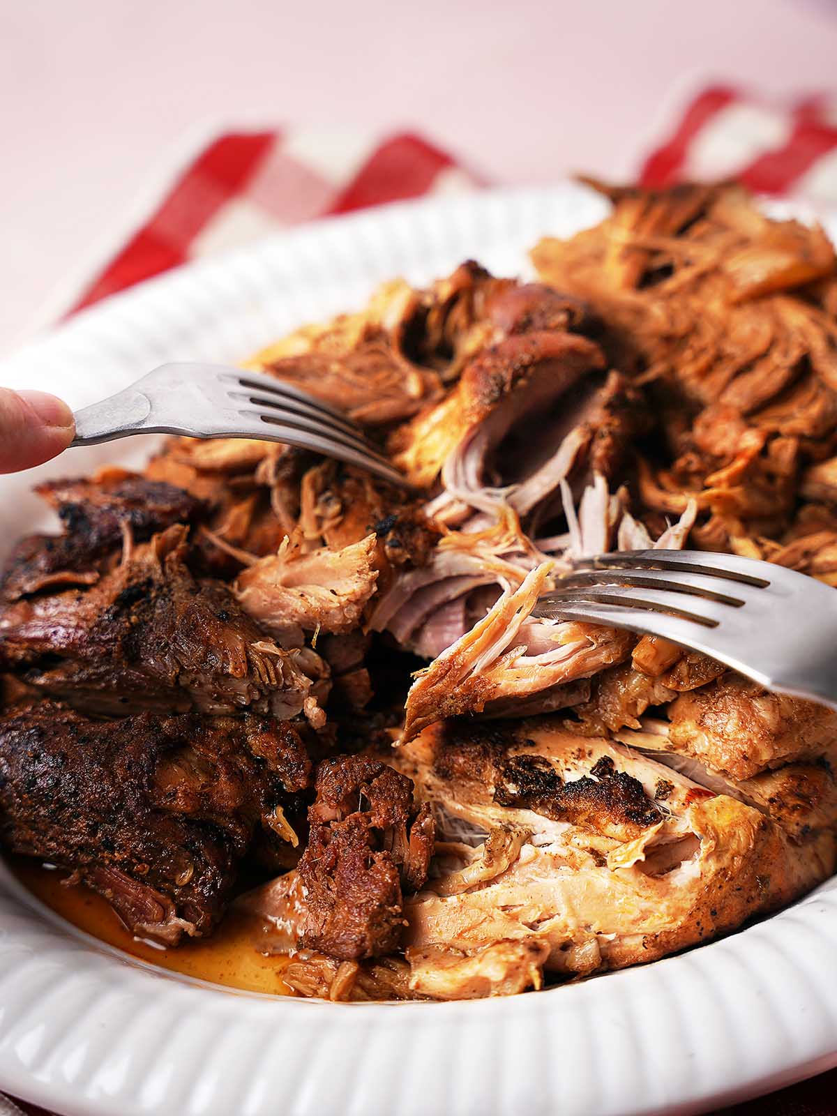 Two forks are shredding tender, seasoned pulled pork on a white plate, with a red and white cloth napkin in the background. The pork looks juicy and well-cooked, with crispy edges visible.