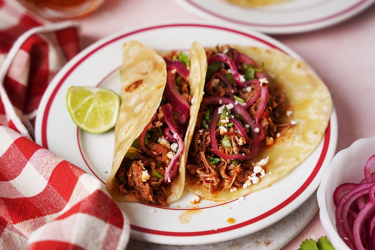 Two soft tacos filled with shredded meat, pickled red onions, cilantro, and crumbled cheese sit on a plate with a lime wedge, next to a red and white checkered napkin.