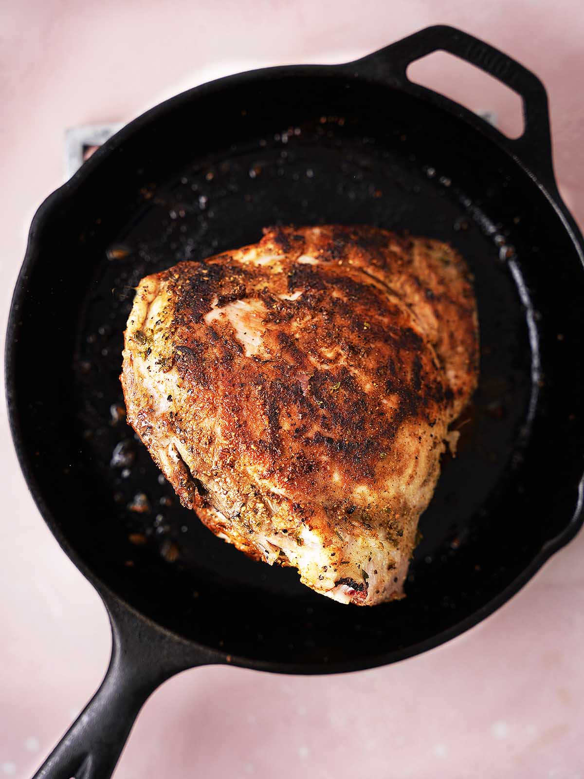 A seasoned, cooked turkey breast sits in a black cast iron skillet, viewed from above against a light pink background.