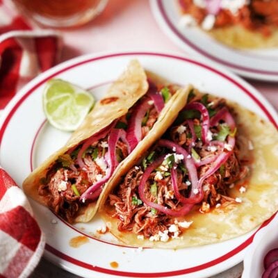 Two pulled pork tacos filled with shredded meat, pickled red onions, cheese, and cilantro sit on a white plate with a red rim, garnished with lime wedges and placed next to a red and white checkered napkin.