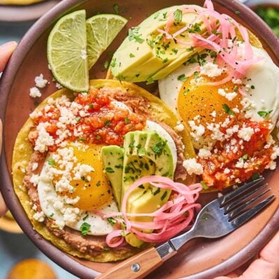 A plate of huevos rancheros with two fried eggs on tortillas, topped with salsa, avocado slices, pickled onions, crumbled cheese, and fresh herbs, garnished with lime wedges. A fork rests on the plate.