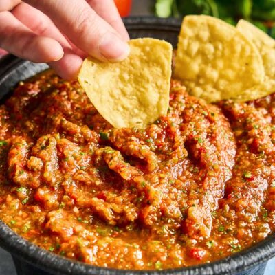 A hand dips a tortilla chip into a bowl of chunky red salsa, with more chips and fresh herbs visible in the background.