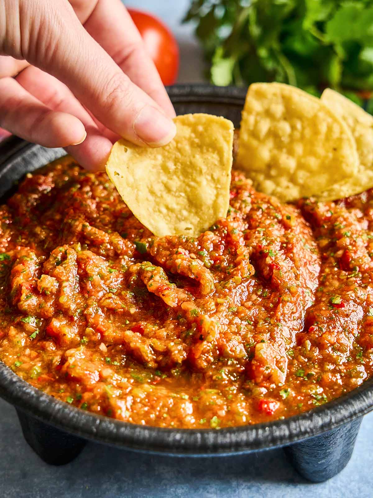 A hand dips a tortilla chip into a bowl of chunky red salsa, with more chips and fresh herbs visible in the background.