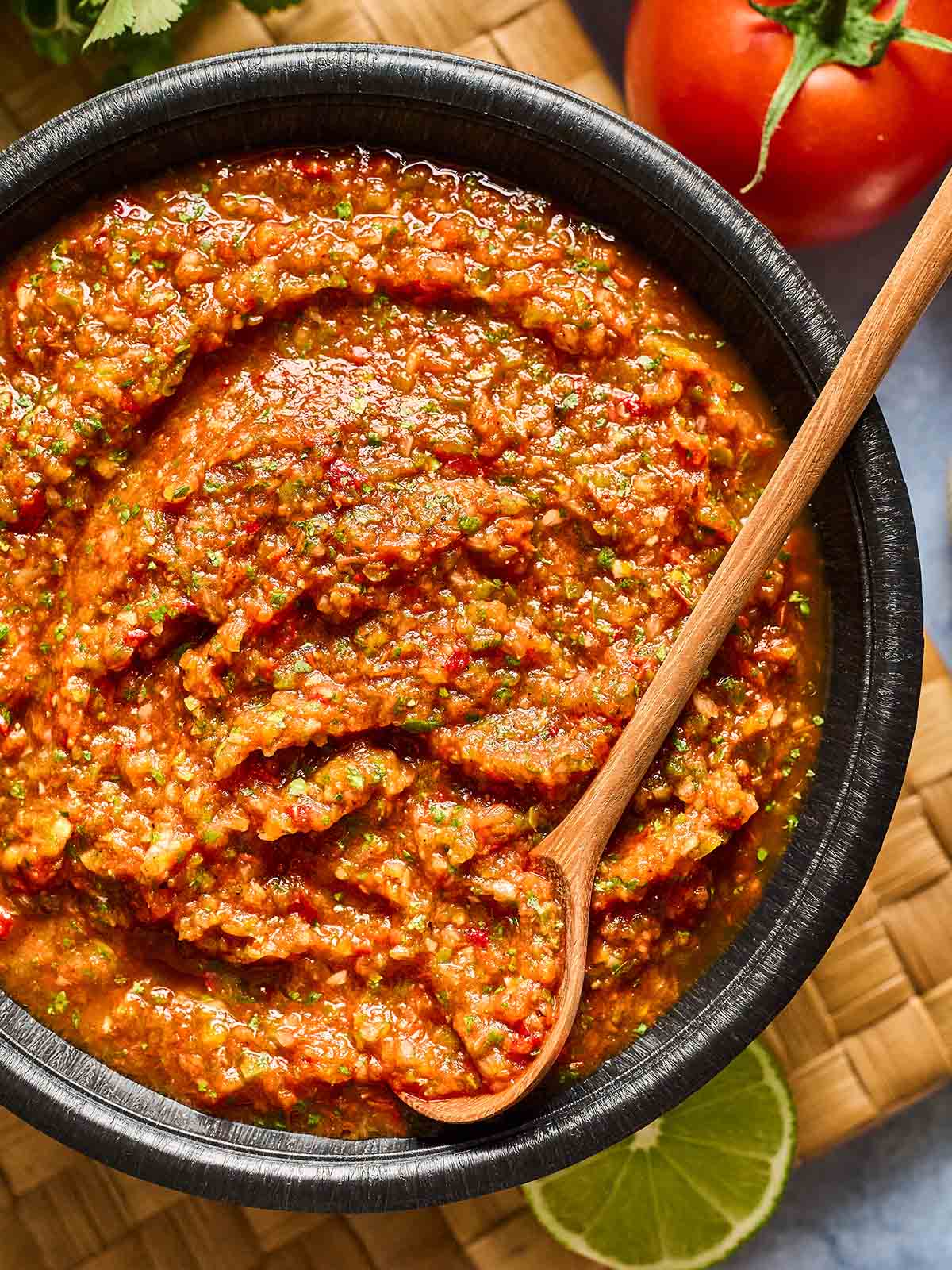 A black bowl filled with chunky red salsa, with visible pieces of tomato, herbs, and spices, and a wooden spoon resting in the salsa. A fresh tomato and a lime slice are nearby on a woven mat.