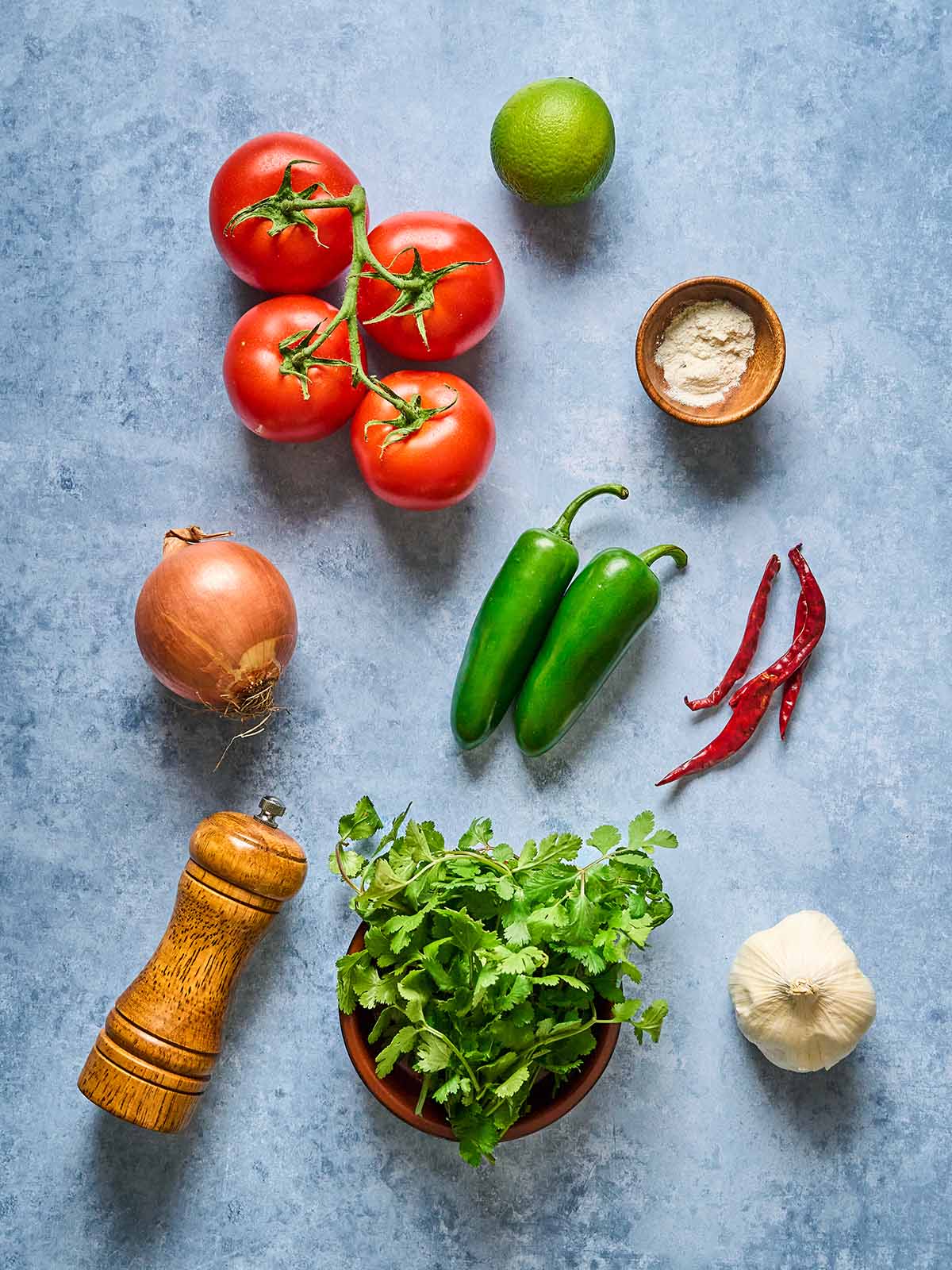 Fresh ingredients on a blue surface: vine tomatoes, a lime, a bowl of flour, two green jalapeños, two dried red chilies, an onion, a wooden pepper grinder, a bunch of cilantro, and a garlic bulb.