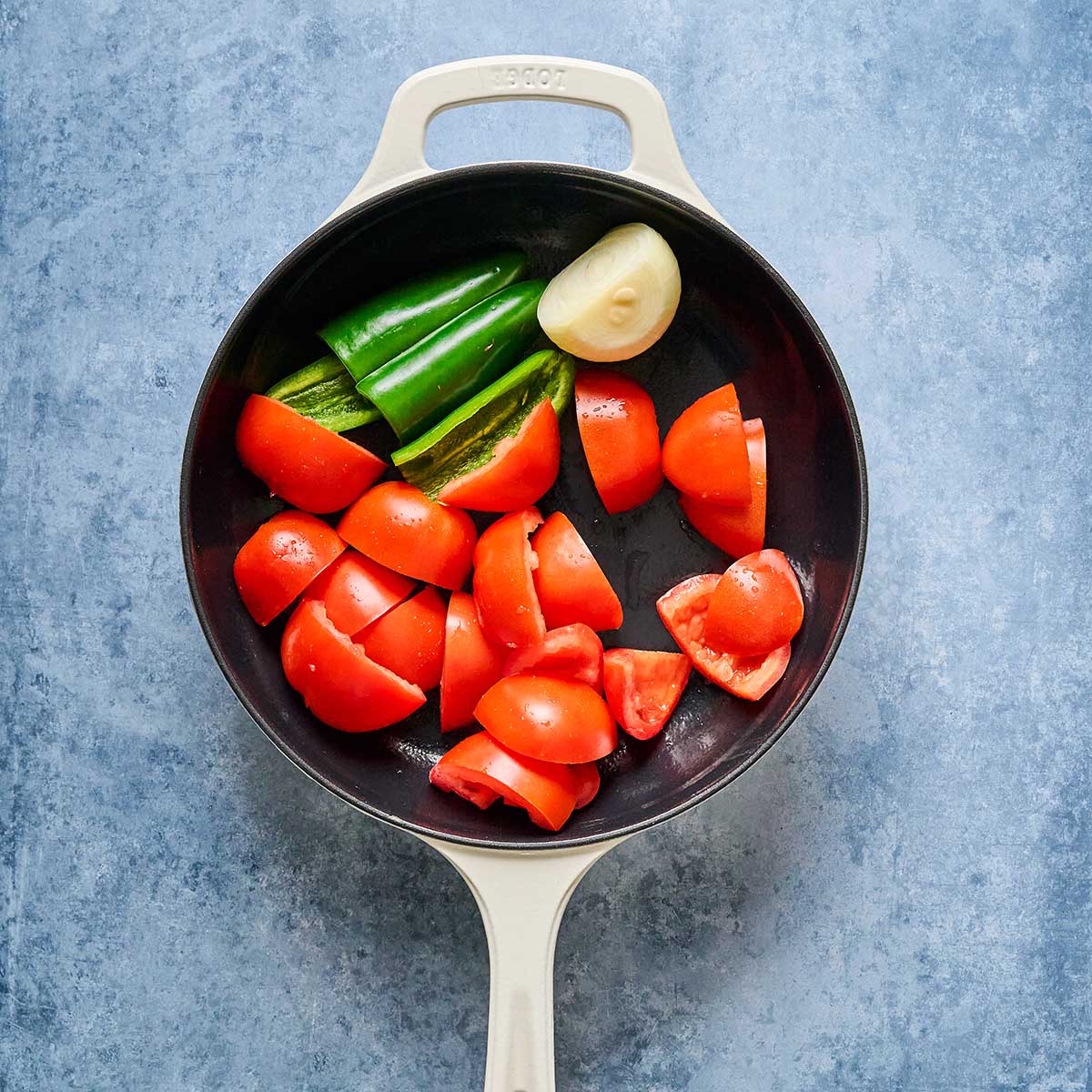 A cream-colored skillet on a blue surface contains chopped tomatoes, halved green jalapeños, and a peeled garlic clove.