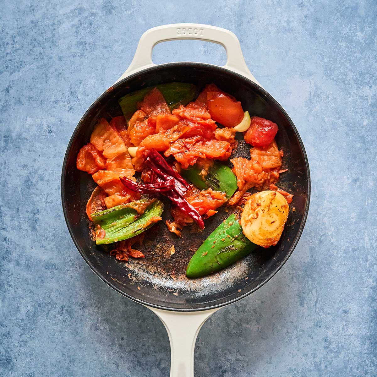 A white skillet on a blue surface contains cooked tomatoes, green peppers, dried red chilies, onion, and garlic cloves.