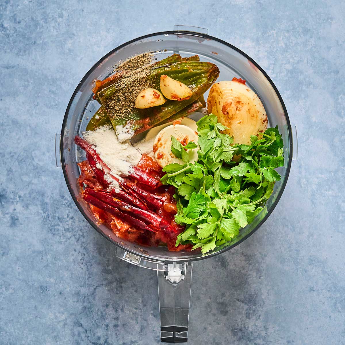 A food processor bowl filled with cilantro, dried red chilies, peeled onion, garlic cloves, green jalapeño, spices, and seasonings, all ready to be blended, on a blue countertop.