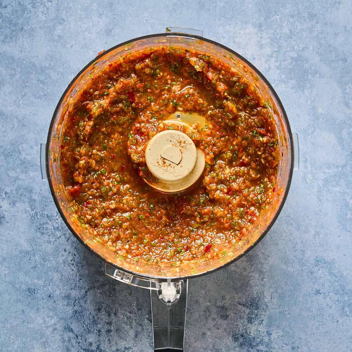 A food processor containing a chunky, reddish-brown salsa with visible pieces of herbs and vegetables, viewed from above on a textured blue surface.