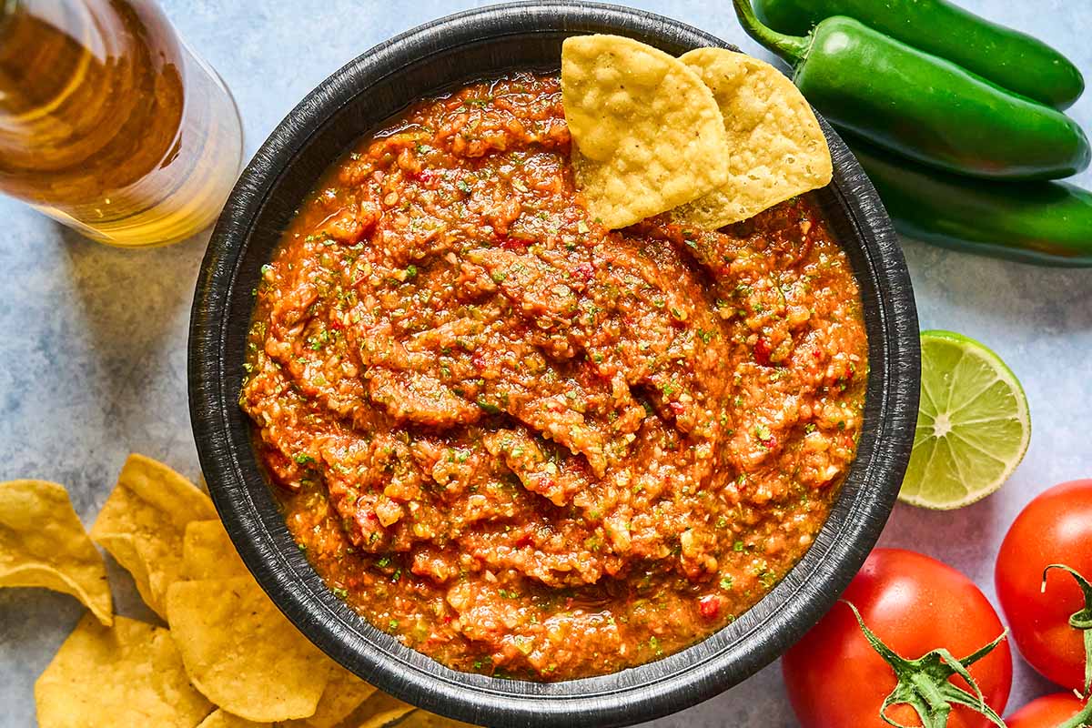 A black bowl filled with chunky red salsa, garnished with two tortilla chips. Surrounding the bowl are whole tomatoes, a lime half, green chili peppers, a bottle, and scattered tortilla chips.