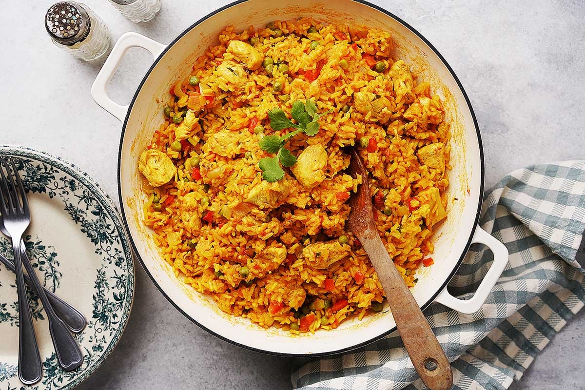 A white pot filled with yellow rice, chunks of chicken, and vegetables, garnished with cilantro, sits on a gray surface beside a checkered cloth, vintage plates, and cutlery.